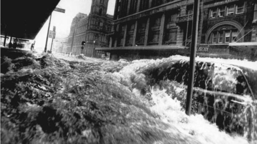 Black and white image of high flood waters rushing down a city street