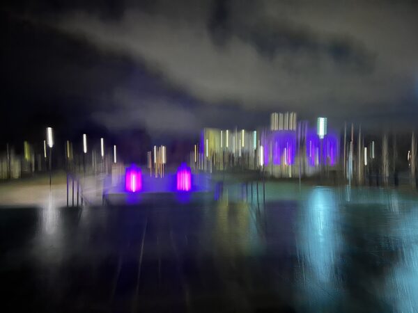 A blurred image of coloured lights at night with clouds in the sky and light staining the dark footpath.